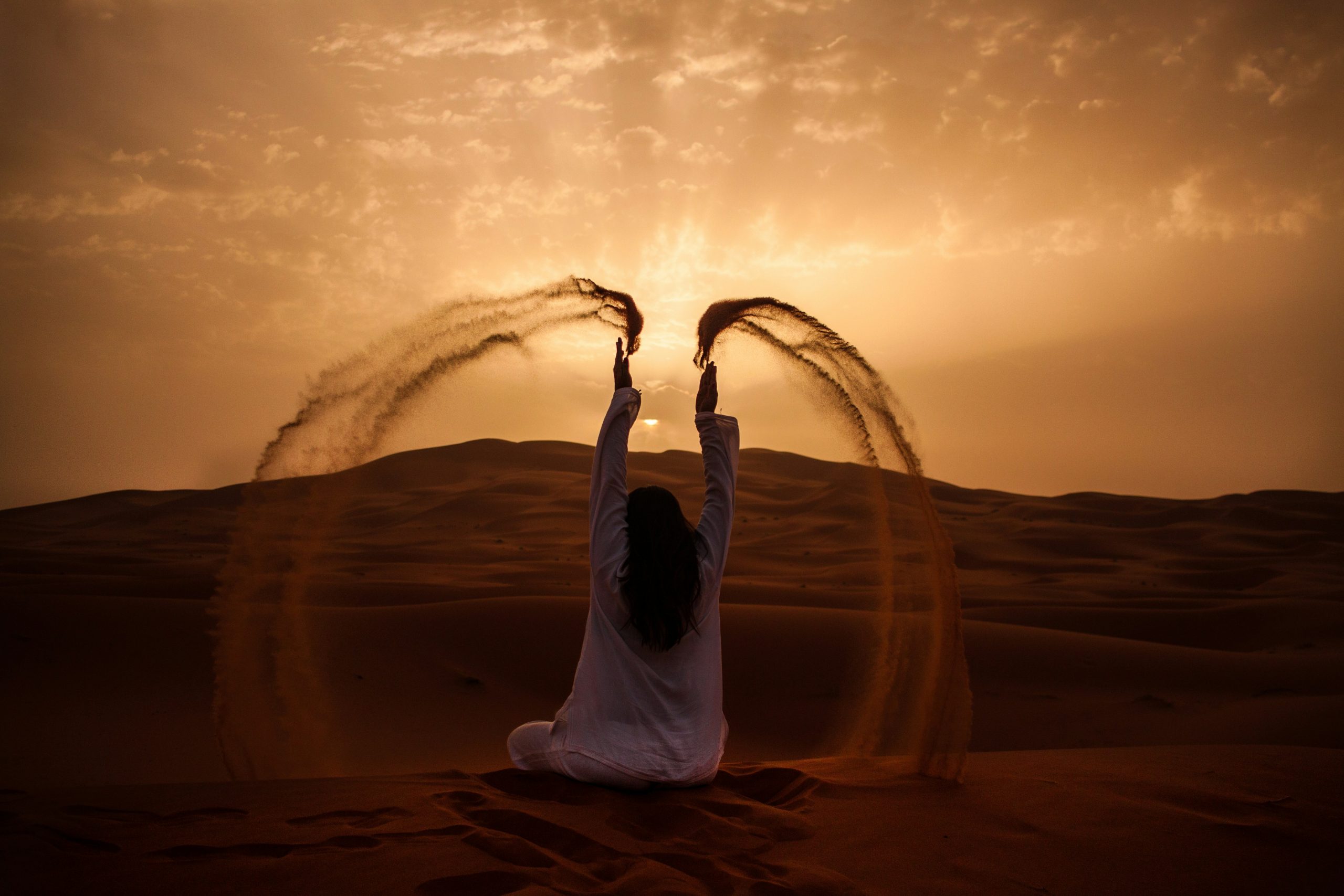 femme assise sur le sable du désert marocain, jetant le sable au dessus de sa tête face à un soleil couchant.