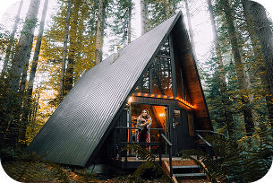 petite maison entourée par la forêt illuminée par des lampes sur sa façade avec une femme sur le porche profitant d'un trek.