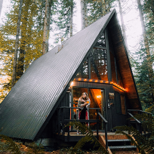 petite maison entourée par la forêt illuminée par des lampes sur sa façade avec une femme sur le porche profitant d'un trek.